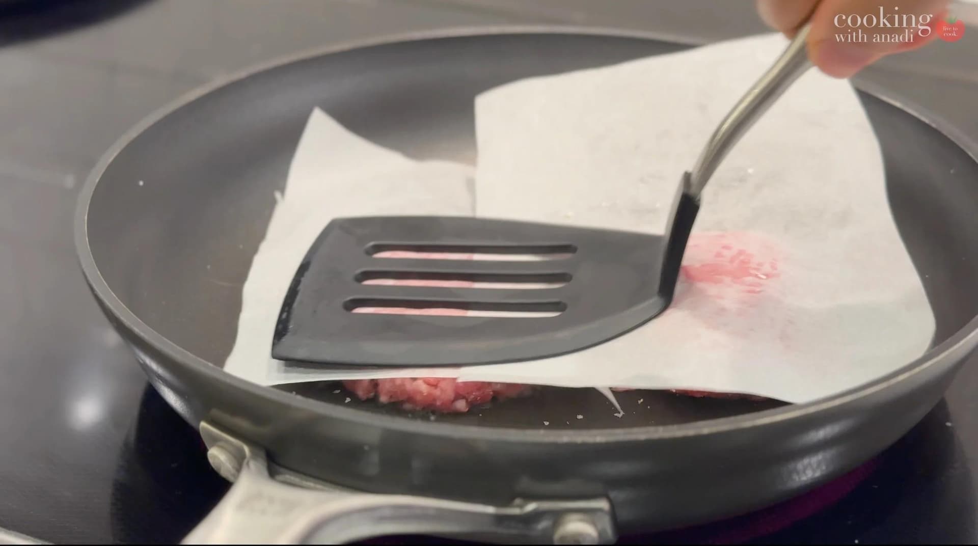 Flattened burger patty being pressed with a spatula in a skillet with parchment paper.
