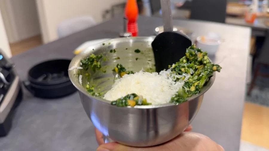 Stainless steel bowl with spinach pesto mixture, grated Parmesan cheese, and a black spoon for mixing.