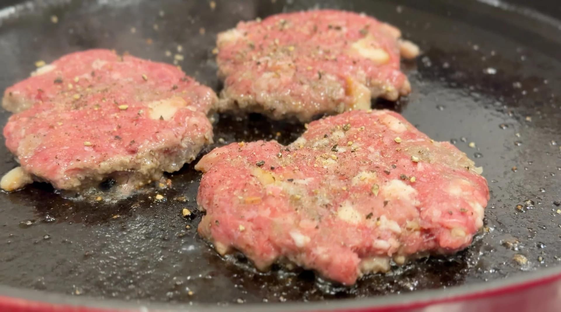Raw burger patties sizzling in a cast iron pan, seasoned with salt and pepper.