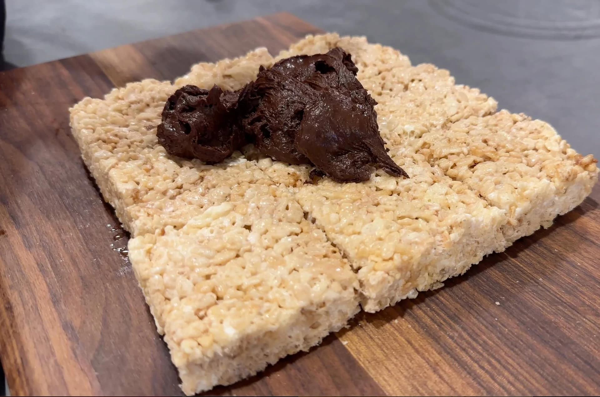 Rice Krispies treat squares cut into portions with chocolate topping spread on one piece, displayed on a wooden board.