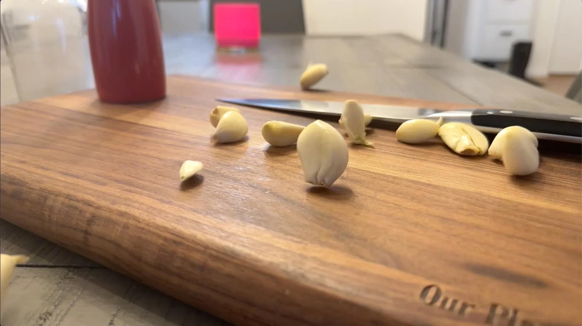 Smashed and peeled garlic cloves scattered on a wooden cutting board next to a chef's knife.