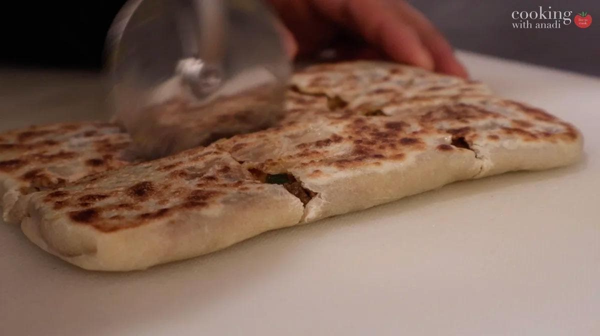 Slicing cooked beef martabak into square pieces with a knife on a white surface.