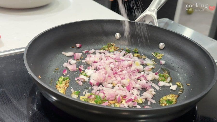 Diced pink onions and green herbs sautéing in a dark skillet with oil and spices.