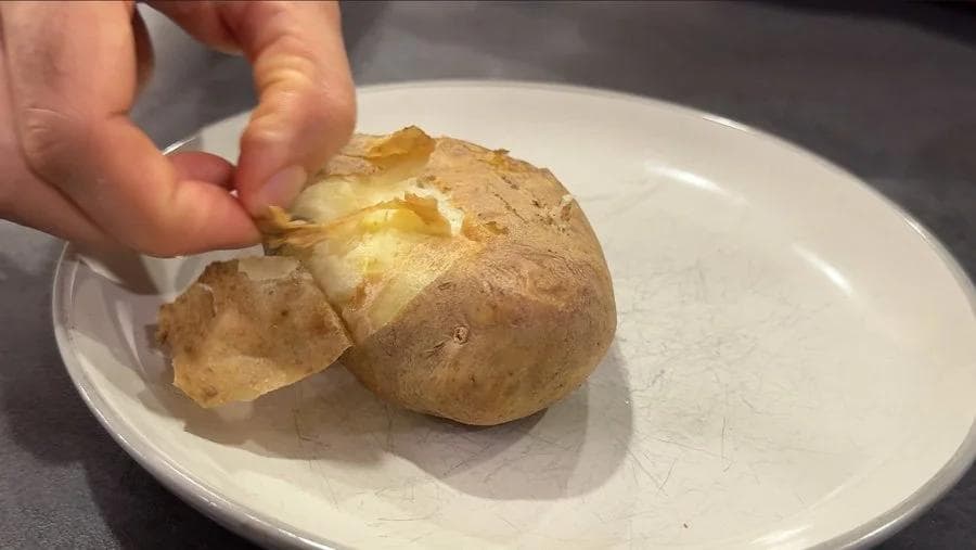 Hand peeling crispy skin off a soft air-fried baked potato on a white plate