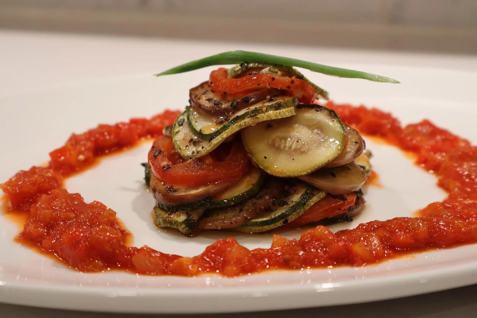 Stacked ratatouille tower with zucchini, tomato, and eggplant slices over chunky tomato sauce, garnished with a green onion.