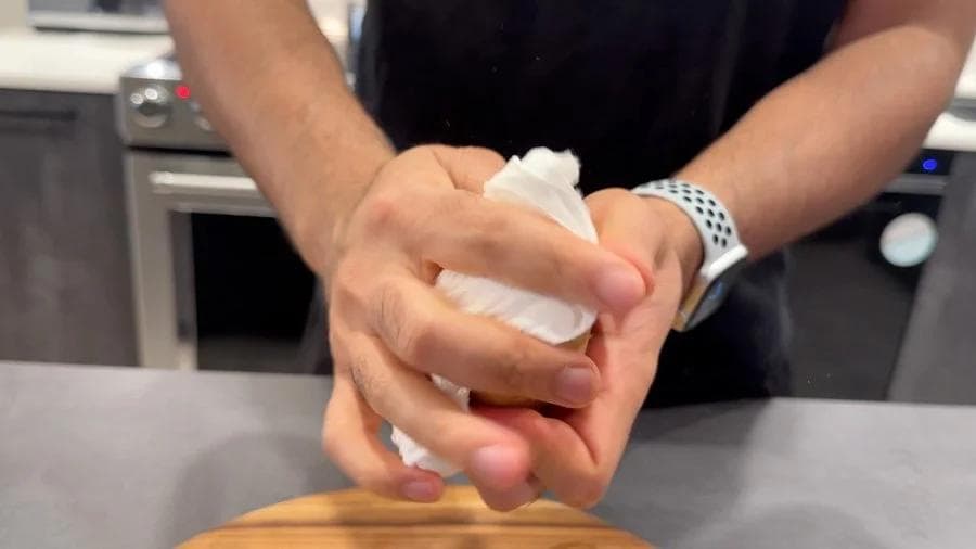 Hands patting a potato dry with a white paper towel over a wooden surface in a kitchen.