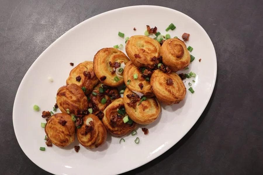 Golden-brown mashed potato pinwheels garnished with crispy bacon bits and chopped green onions on a white plate.