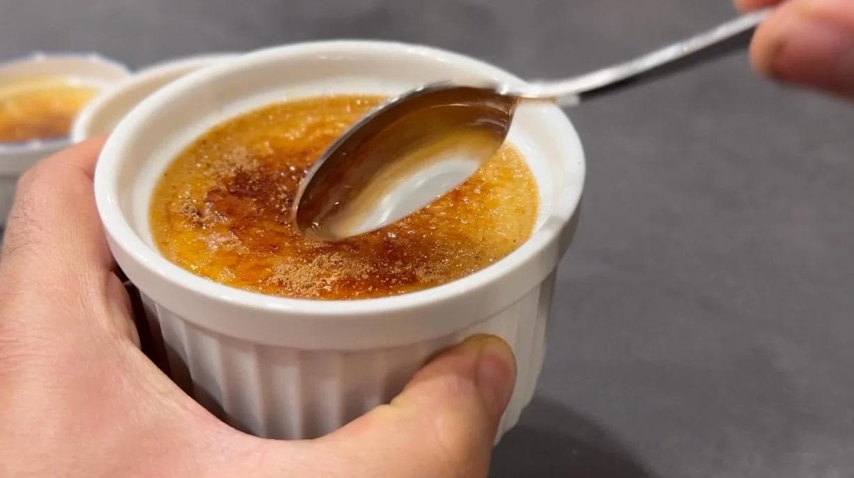 Hand holding a white ramekin of creme brulee with a spoon cracking through the caramelized sugar top.