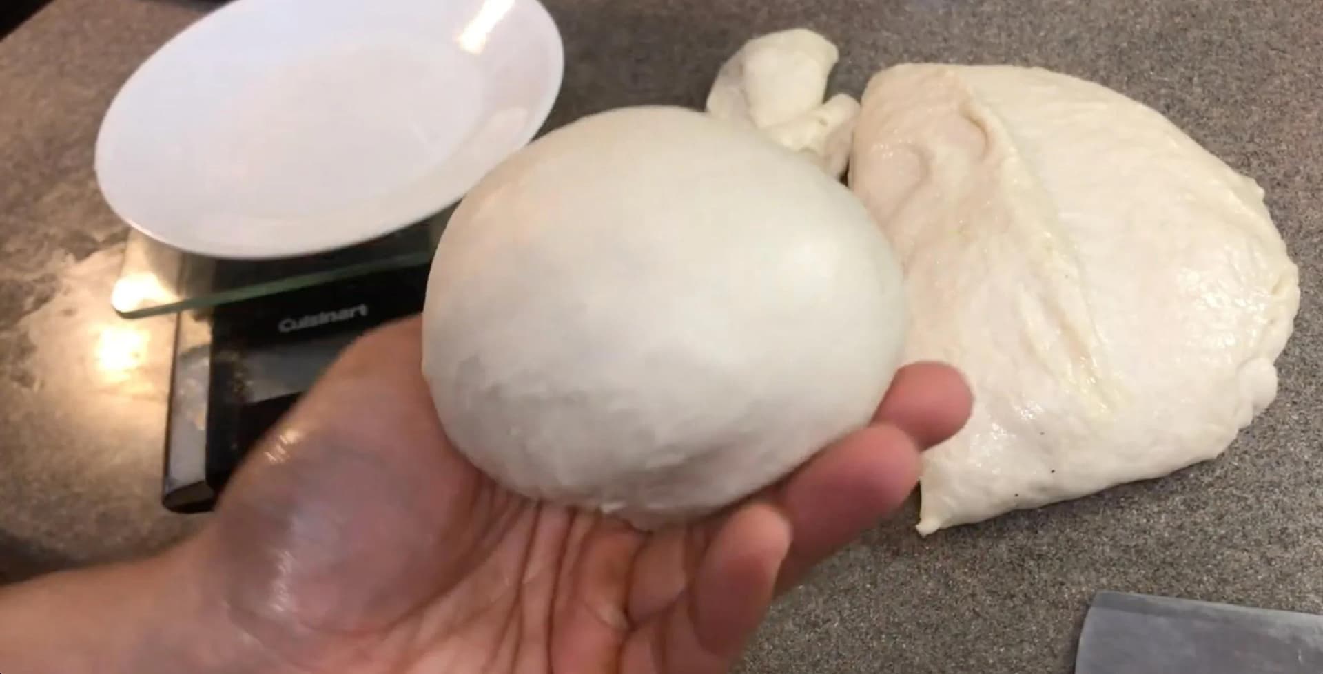 Hand holding a round ball of white dough next to a kitchen scale and white plate.