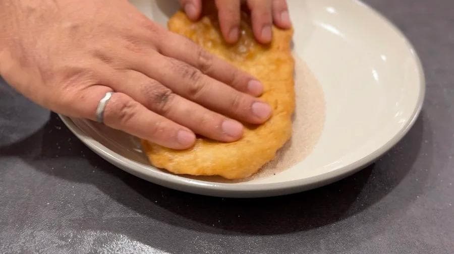 Hand pressing a fried pastry into cinnamon sugar on a white plate.
