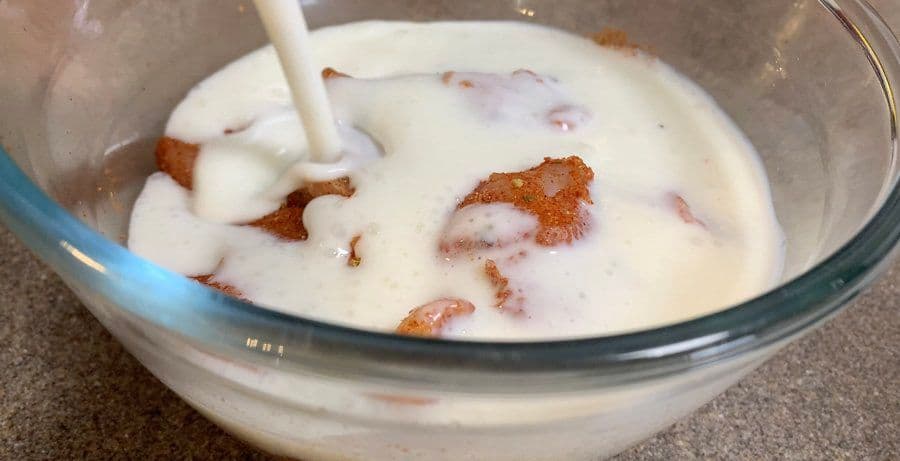 Cubed chicken coated in spice mix being submerged in buttermilk in a glass bowl.
