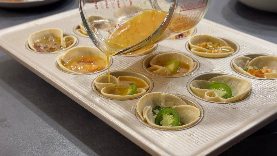 Pouring broth into baked egg cups topped with jalapeños and cheese in a muffin tin.