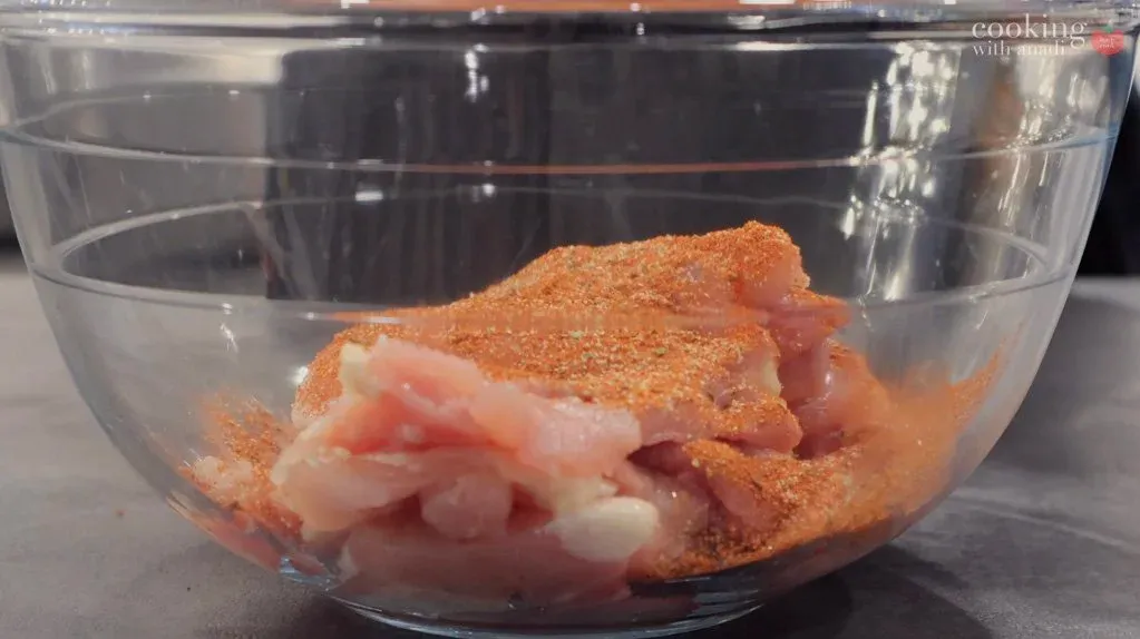 Raw chicken pieces coated in orange spice mixture in a glass bowl.