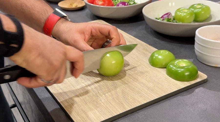 Hands cutting whole green tomatillos in half on a wooden cutting board for salsa verde.