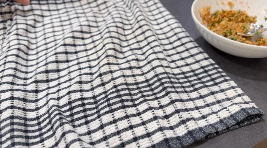 Blue and white checkered kitchen towel on gray surface with a bowl of cauliflower mixture nearby.