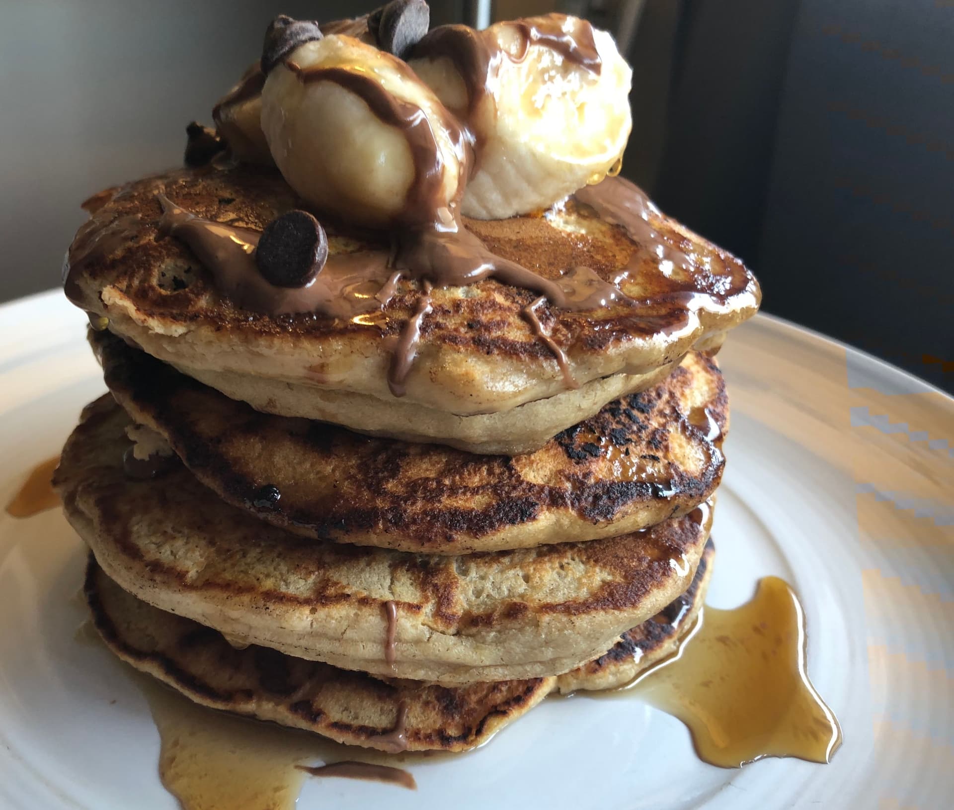 Stack of four fluffy pancakes topped with whipped cream, chocolate drizzle, and chocolate chips, served with maple syrup.