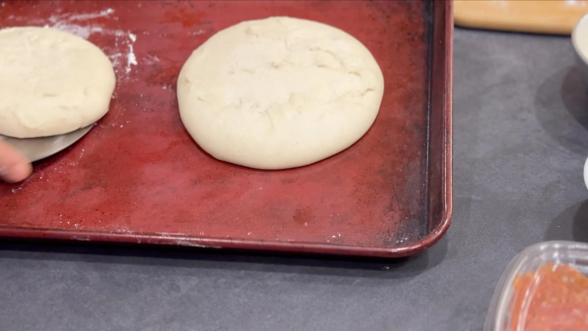 Two pizza dough balls being stretched on a flour-dusted red baking surface