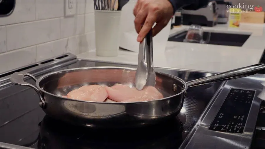 Tongs lifting poached chicken breasts from simmering water in a skillet on an induction cooktop.