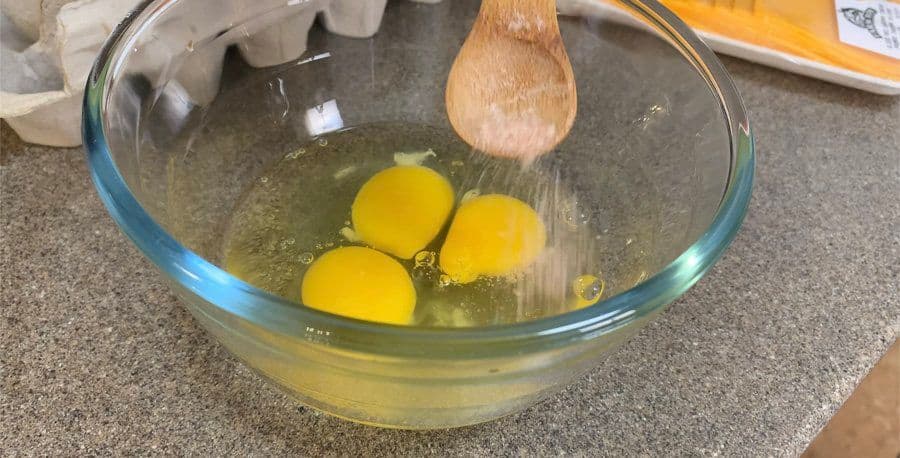 Three eggs whisked with butter in a glass bowl with a wooden spoon, ready for cooking.