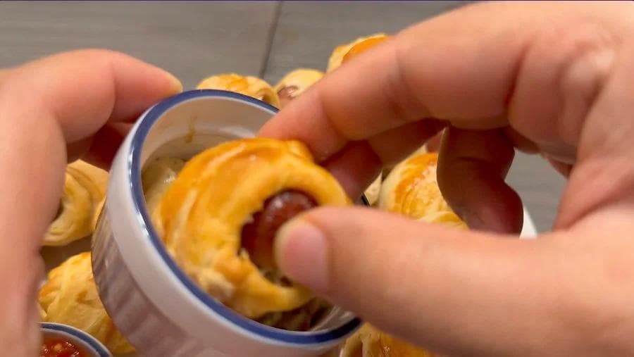 Hand holding a crescent roll pig in a blanket with golden-brown pastry and red sausage visible inside.