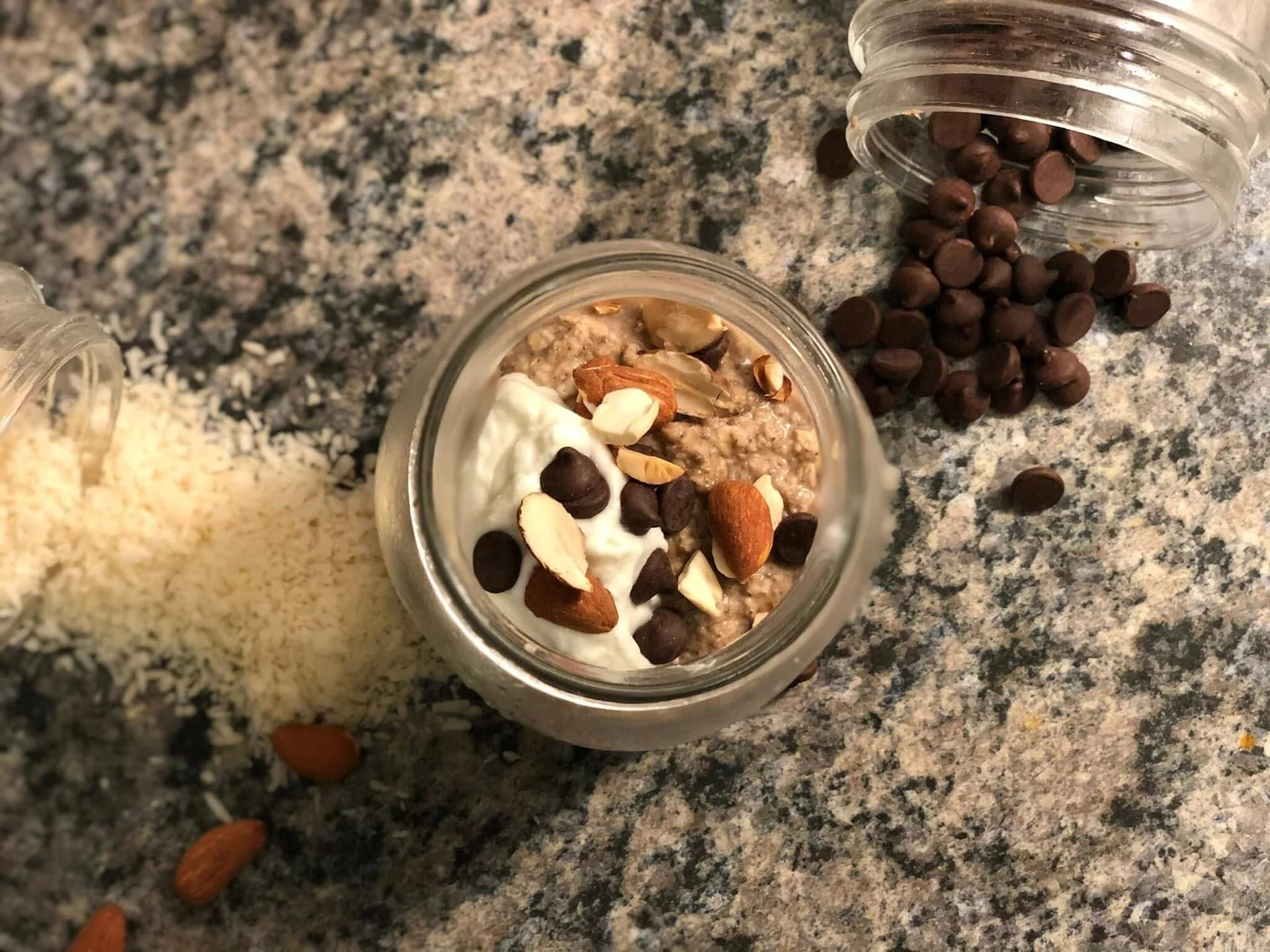Overnight oats in a glass jar topped with whipped cream, chocolate chips, and almonds, with chocolate chips scattered nearby.