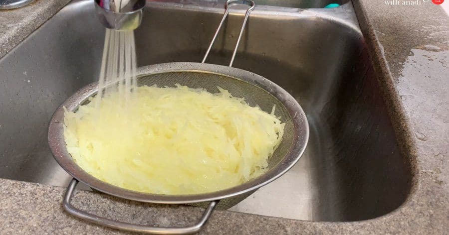 Shredded potatoes being rinsed in a sieve over a kitchen sink with water streaming down.