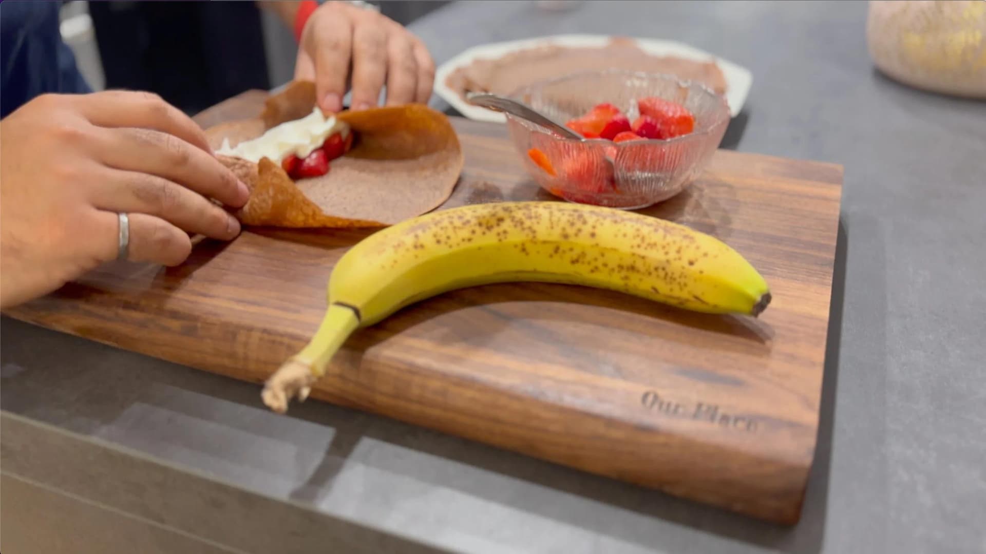 Hands assembling a gluten-free chocolate crepe with sliced strawberries and banana on a wooden cutting board.