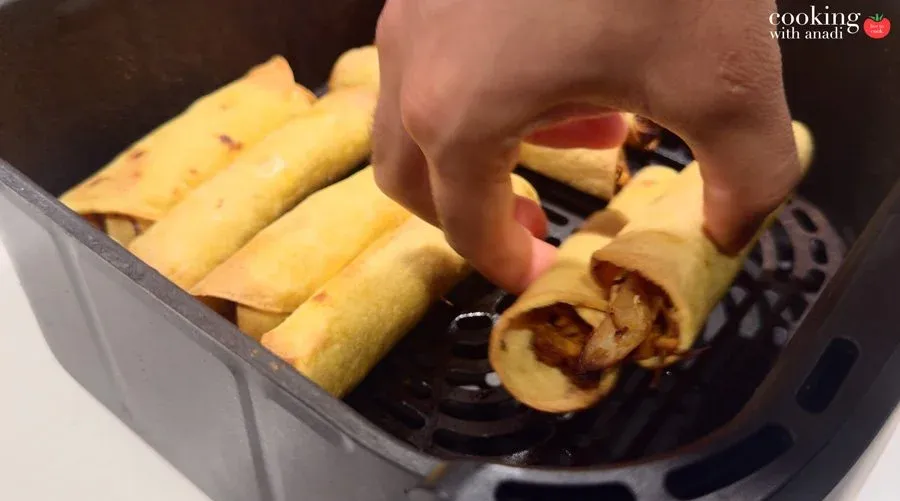 Hand placing rolled taquitos seam-side down into a black air fryer basket with several others already arranged inside.