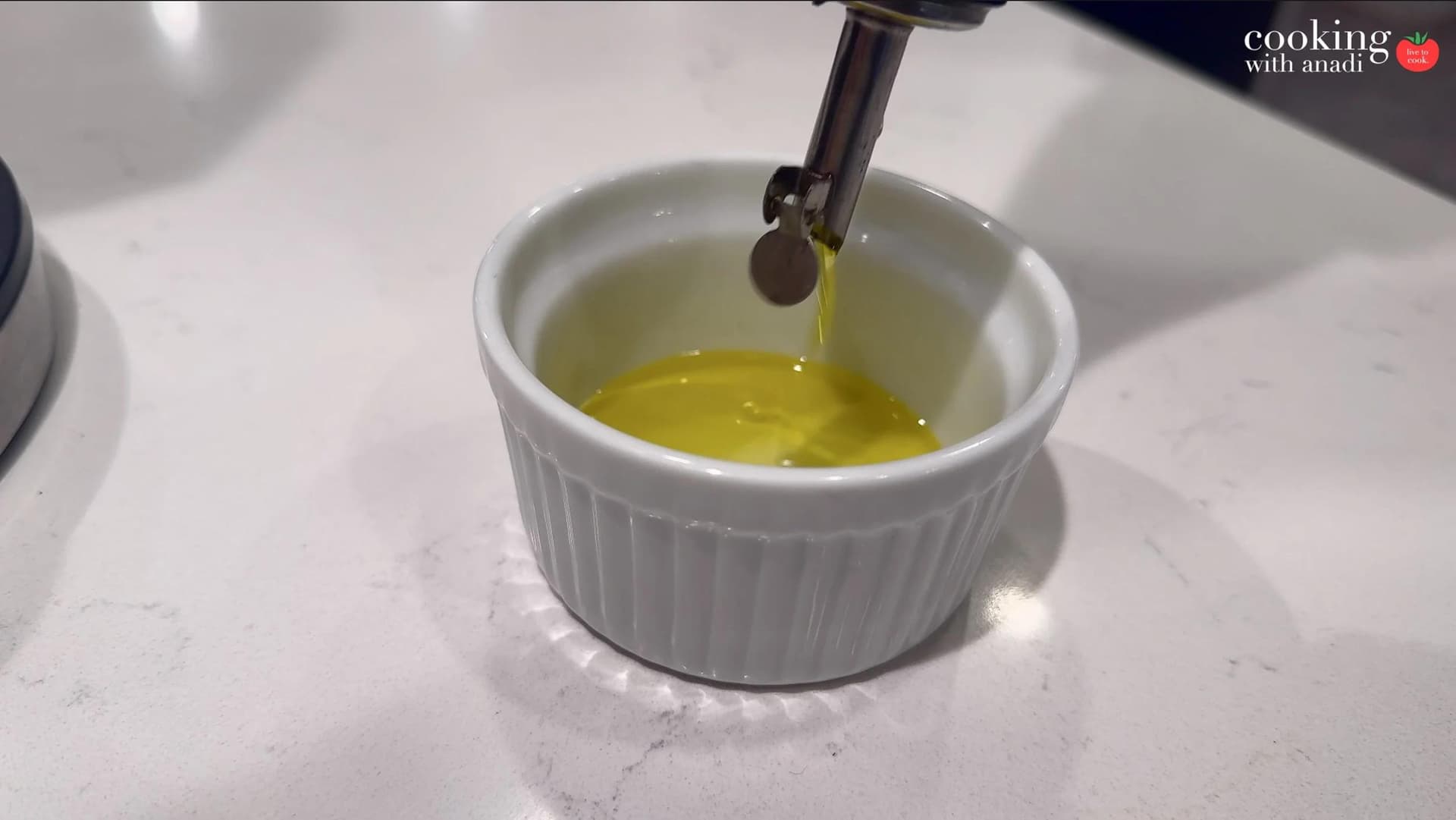 Olive oil being poured from a bottle into a small white ribbed ramekin on a marble surface.