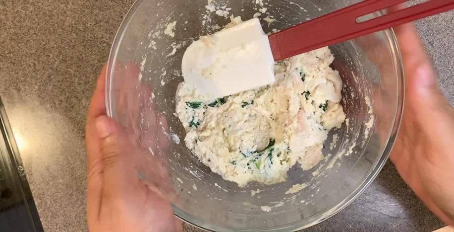 Ricotta cheese mixture with spinach and herbs being mixed in a glass bowl with a red spatula