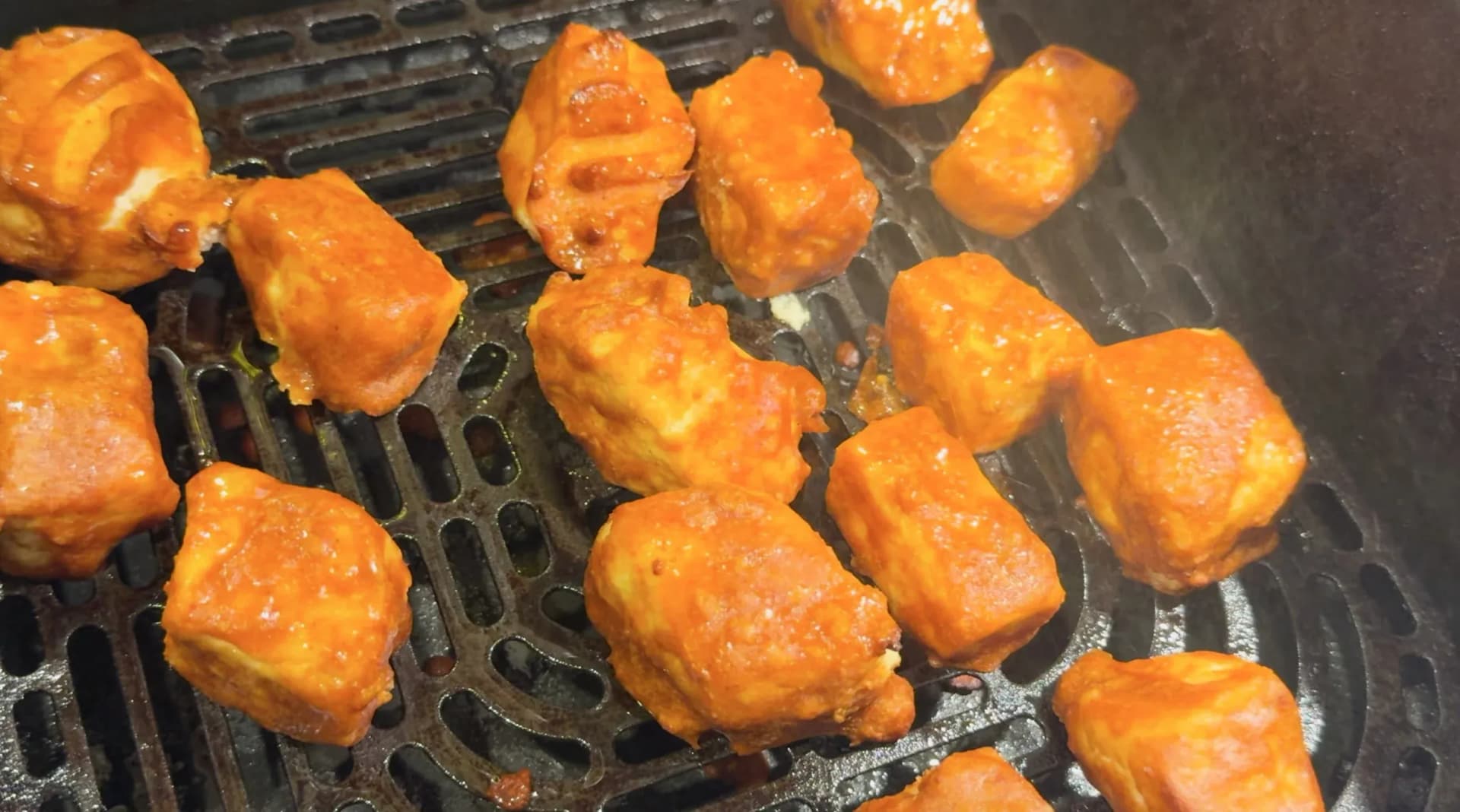Marinated paneer cubes coated in orange-red chilli sauce arranged in an air fryer basket, mid-cook.
