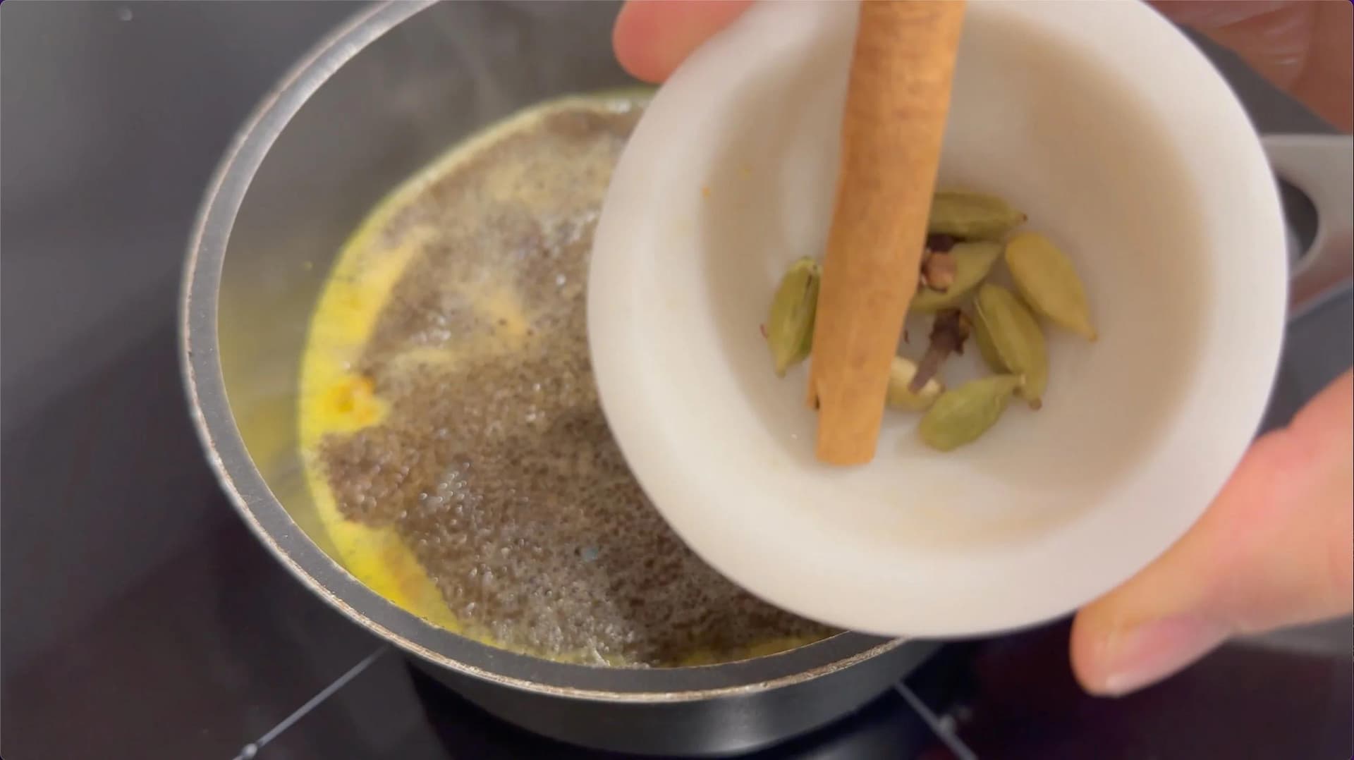 Pouring ground spices from mortar and pestle into simmering turmeric chai base in saucepan.