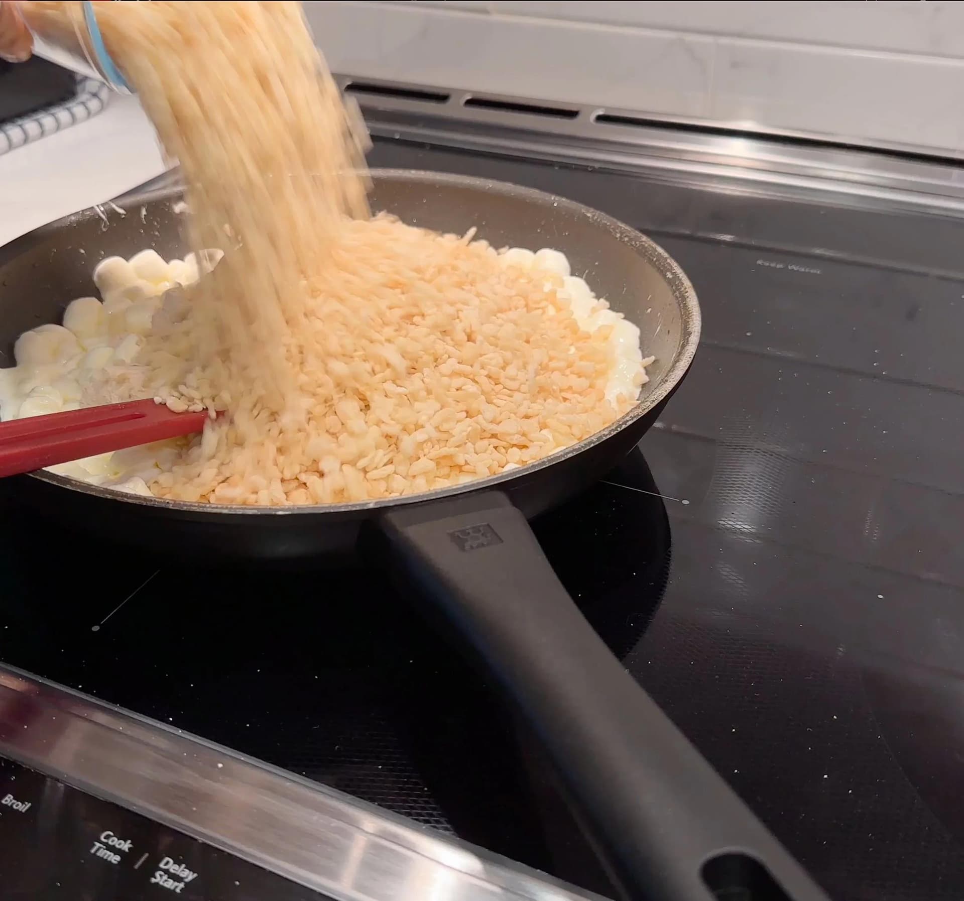 Pouring crisped rice cereal into a pan of melted marshmallows with protein powder on a stovetop.