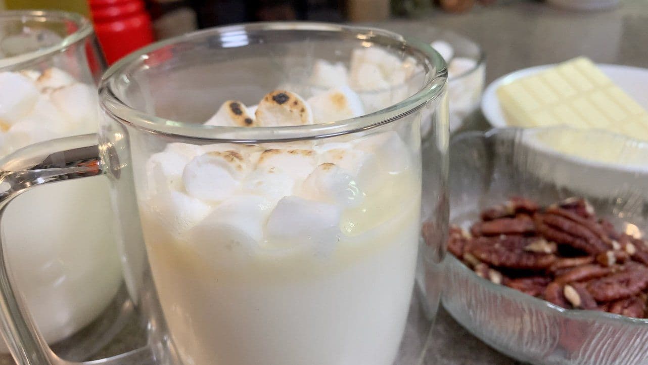 White hot chocolate in a glass mug with whipped cream and chocolate drizzle, surrounded by ingredients like butter and pecans.
