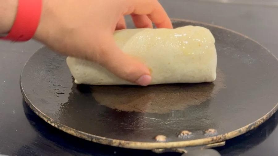 Hand pressing a toasted breakfast burrito onto a dark skillet, showing golden-brown exterior.