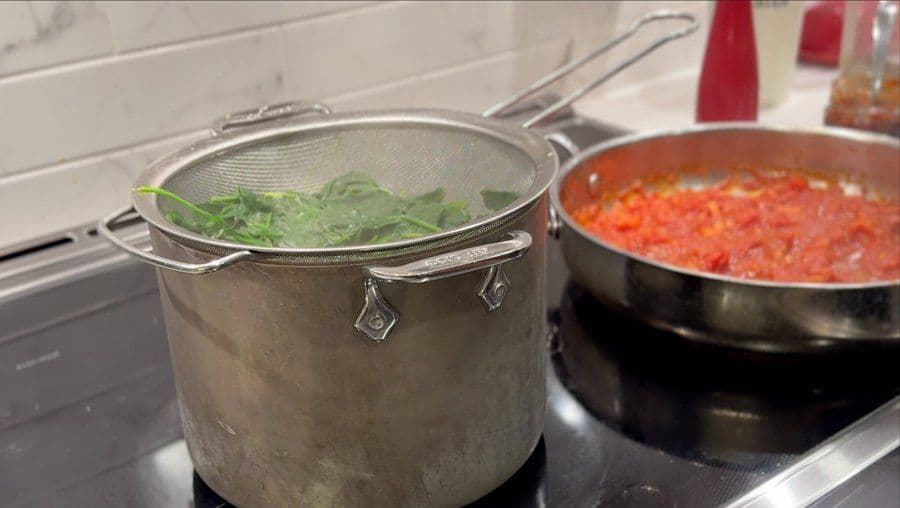 Blanched spinach draining in a colander next to a pot of simmering red Amatriciana sauce on the stovetop.
