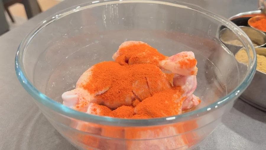 Raw chicken drumsticks coated with red Kashmiri chili powder in a glass bowl during marinating process.