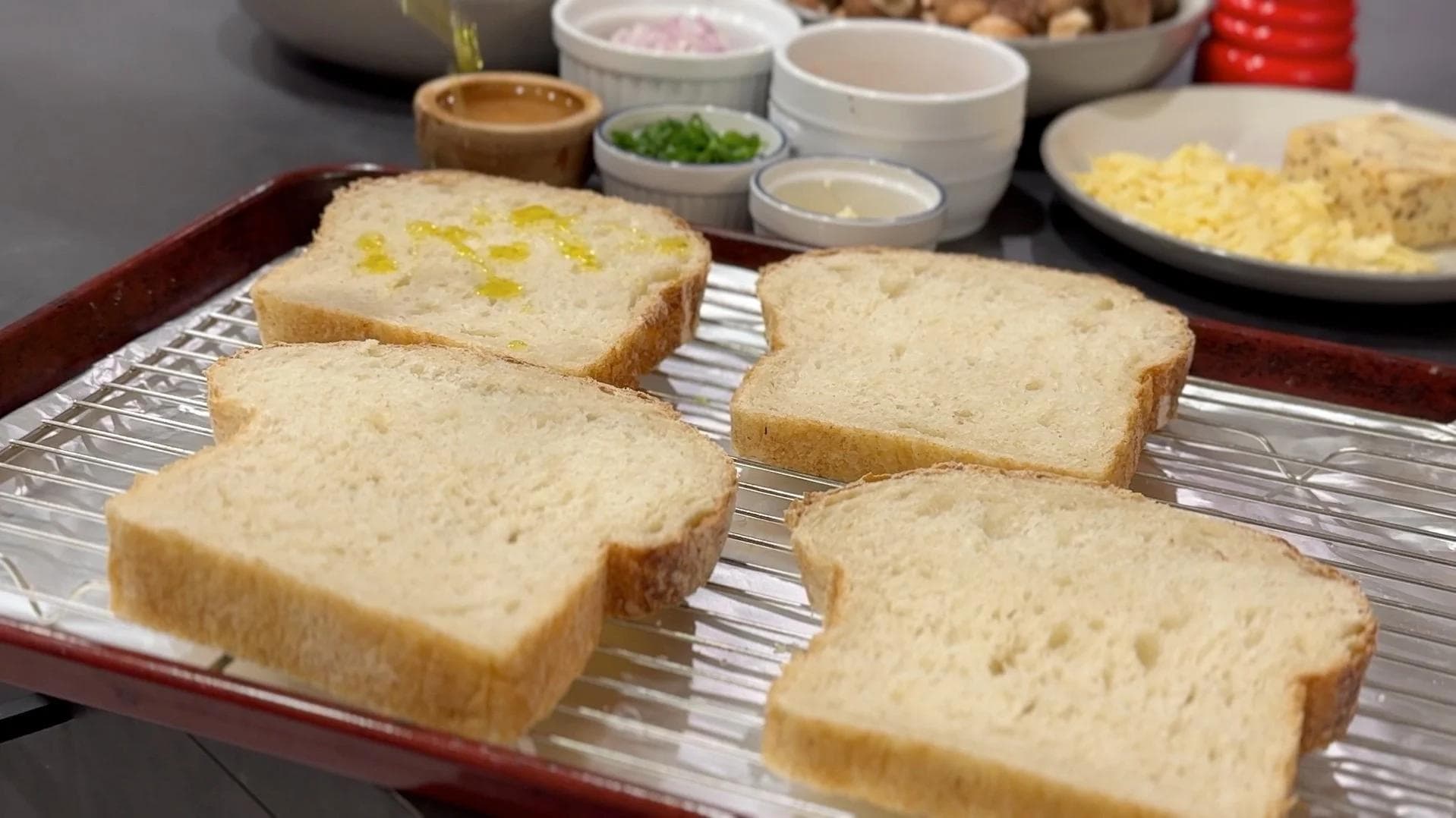 Four slices of toasted sourdough bread on a wire rack, with one slice drizzled with olive oil, surrounded by ingredients in small bowls.