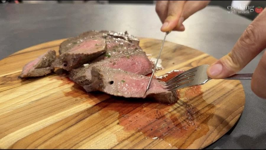 Sliced air fryer steak with pink center being cut with fork, topped with melted herb butter on wooden board.