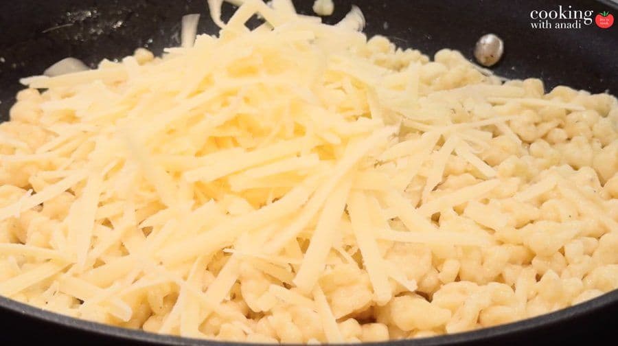 Shredded cheese being folded into cooked spätzle in a dark skillet over medium heat.