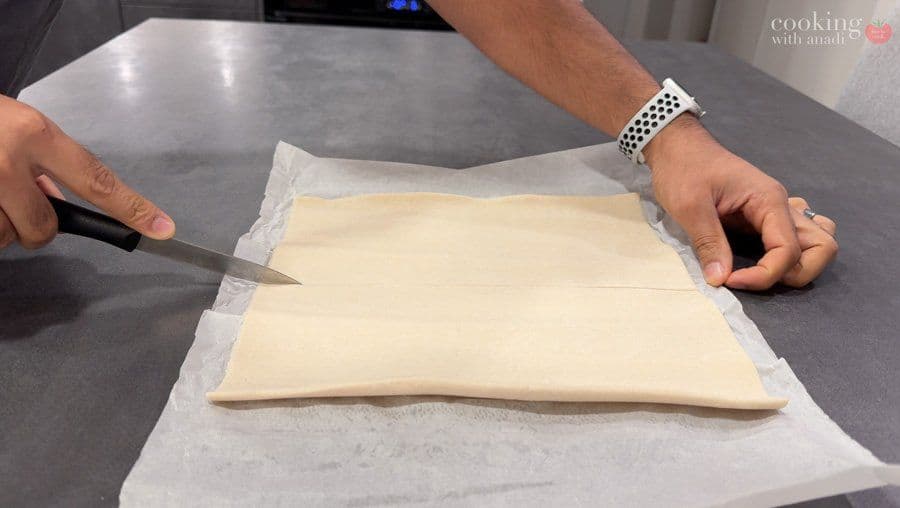 Cutting puff pastry squares on parchment paper with a knife for potato filling.