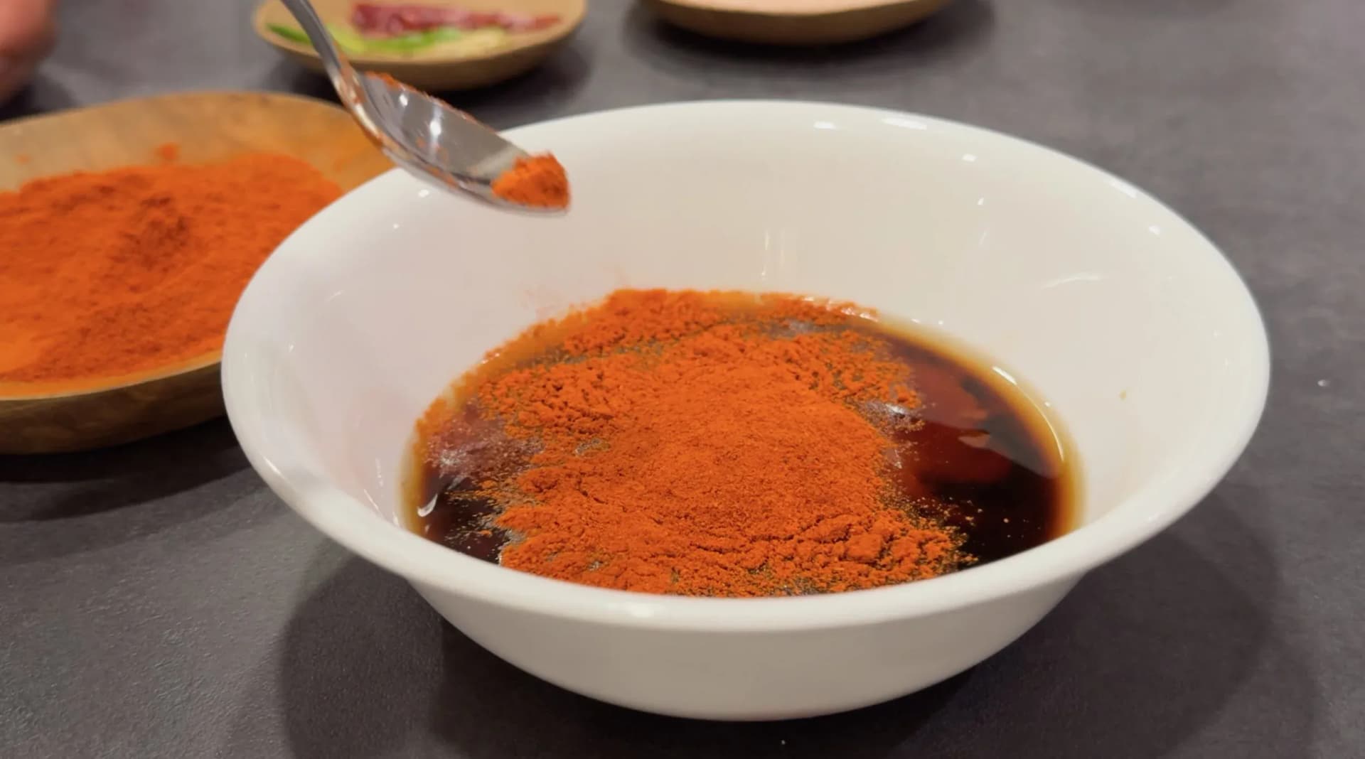Red chilli powder being spooned into a white bowl containing dark soy sauce mixture, with more chilli powder on a wooden plate beside it.