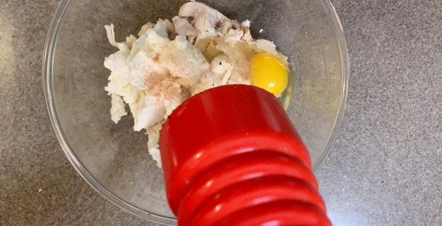 Red spatula mixing ricotta, shredded chicken, and egg in a glass bowl for lasagna filling.