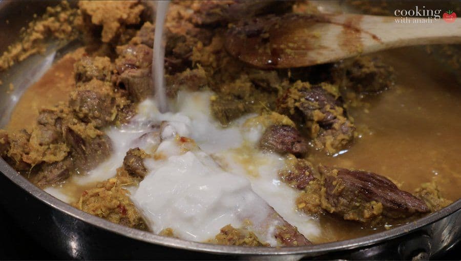 Beef rendang simmering in pan with coconut milk being stirred in, showing browned beef pieces and spice paste.