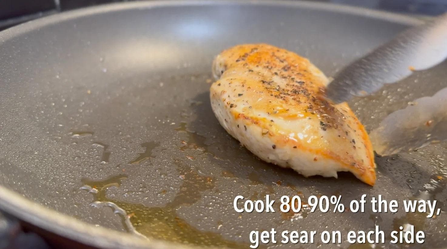 Seared chicken breast in a pan with garlic oil, showing golden-brown crust on top side being flipped with a spatula.
