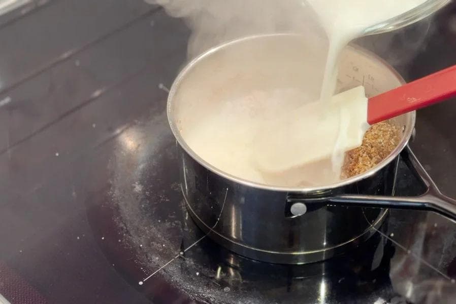 Milk being poured into a pot of roasted cracked wheat with a red spoon stirring the mixture on a stovetop.