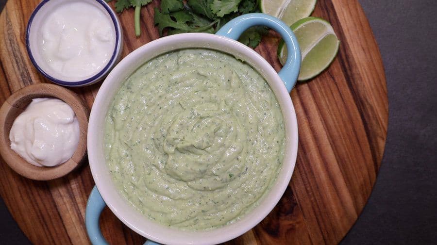 Creamy avocado lime crema in a white bowl with lime wedges, cilantro, and sour cream on the side.