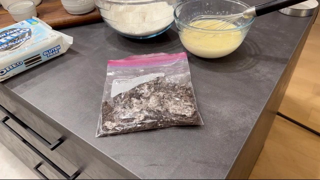 Crushed Oreo cookies in a plastic bag next to bowls of dry and wet pancake batter ingredients on a kitchen counter.