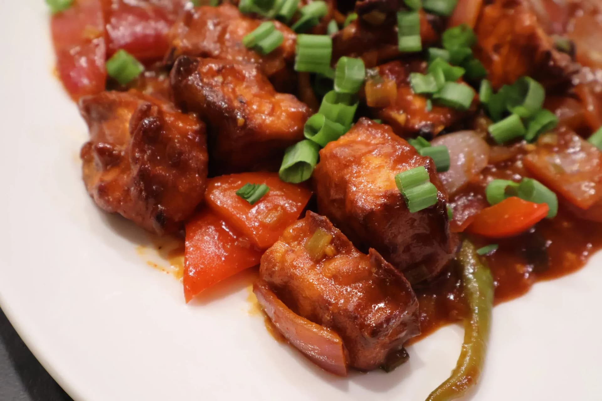 Crispy air fryer chilli paneer cubes in spicy tomato sauce with bell peppers, onions, and chopped green onions on a white plate.