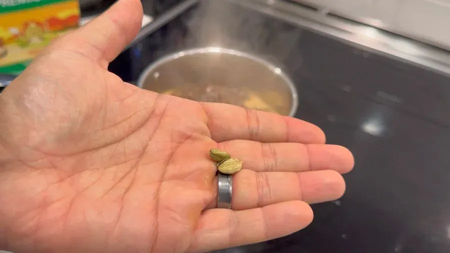Hand holding a lightly smashed cardamom pod over a saucepan of simmering chai tea.